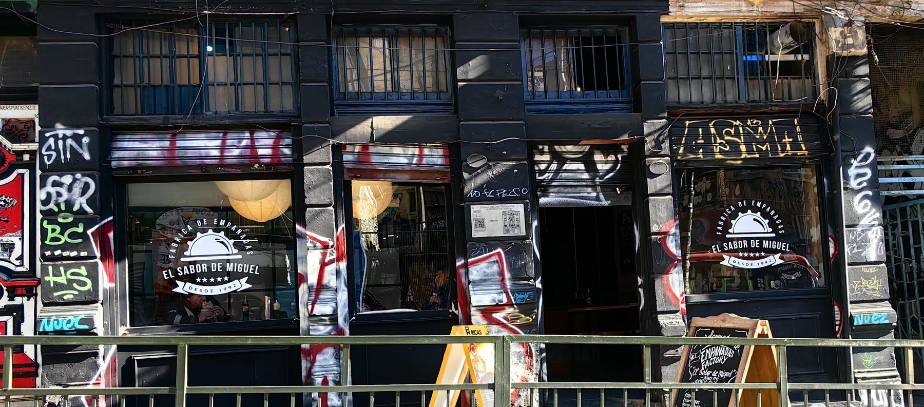 Fachada negra de El Sabor de Miguel en Valparaíso, con el logotipo de la fábrica pintado en las vitrinas y la pizarra de la carta en la vereda.